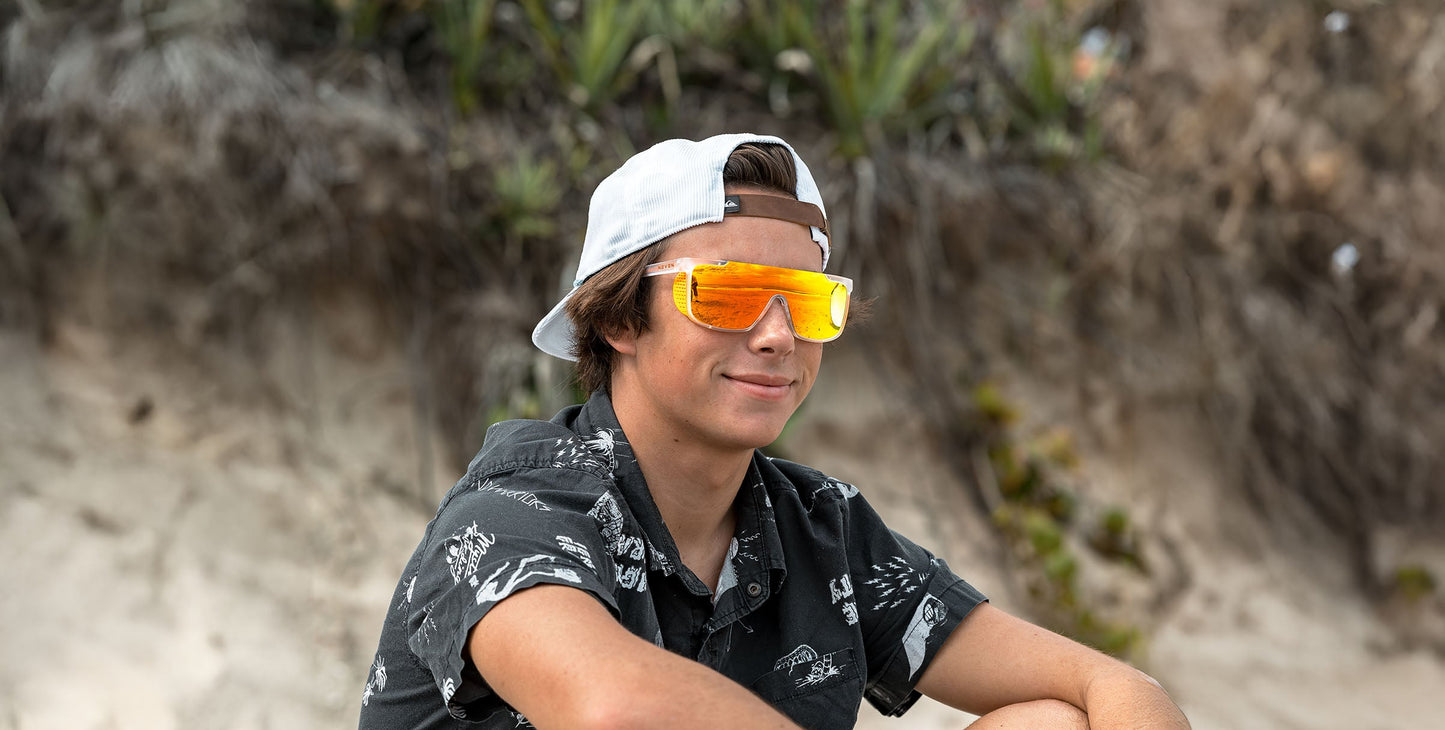 A young person in a white backwards cap and black patterned shirt sits outdoors, smiling slightly while wearing Neven Eyewear's Sunflare orange mirrored polarized sunglasses with UV400 protection.