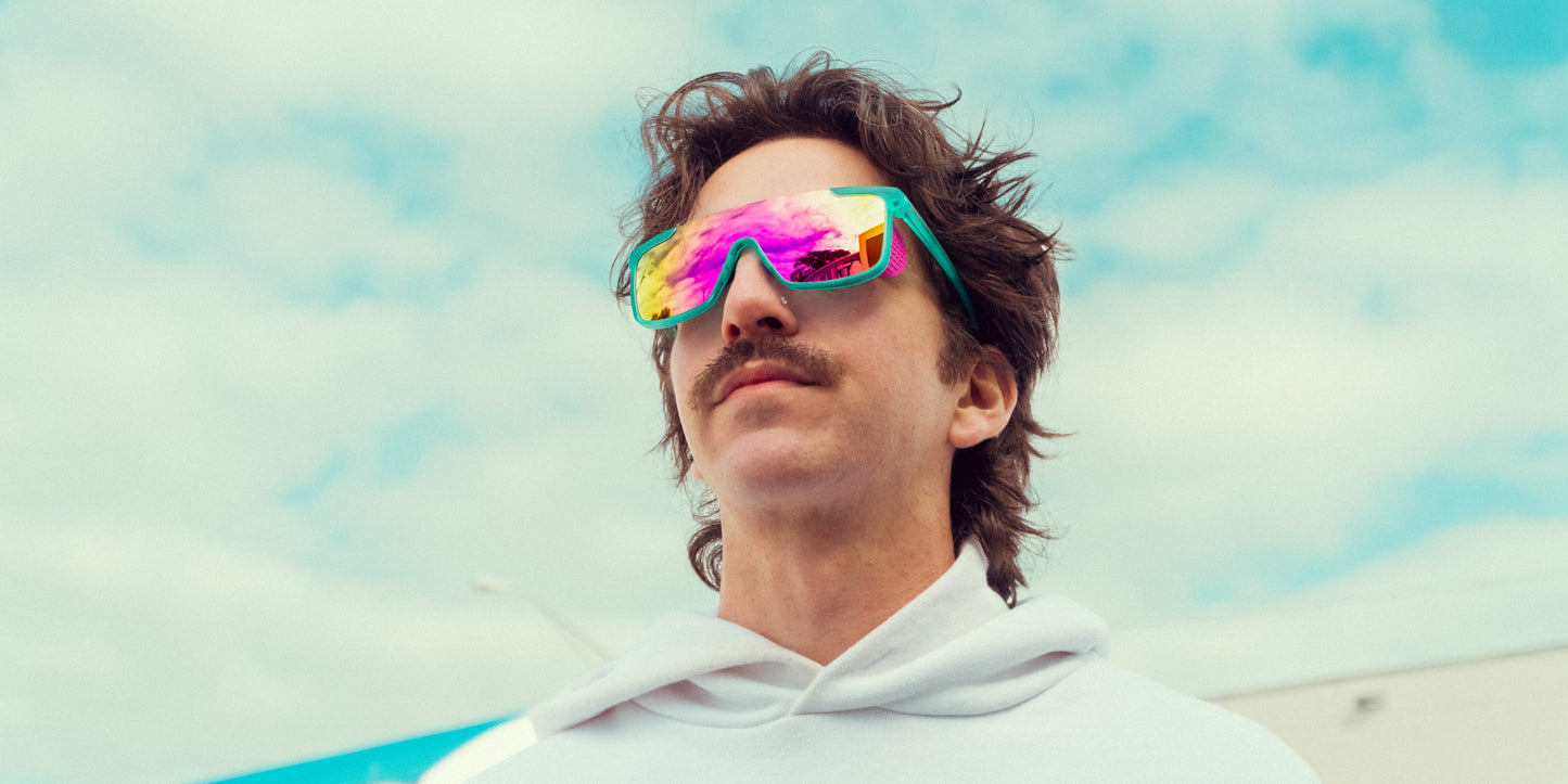 A person with tousled brown hair and a mustache wears a white hoodie and Neven Eyewear's T.D.K. sunglasses with mirrored lenses. The partly cloudy sky and blurry building in the background suggest an outdoor setting.
