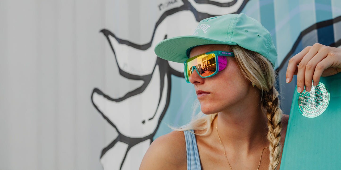 A young woman with long blonde hair wears Neven Eyewear’s T.D.K. polarized mirrored sunglasses and a teal cap, holding a teal skateboard as she stands confidently before a vivid mural of abstract black-and-white shapes and blue tones.