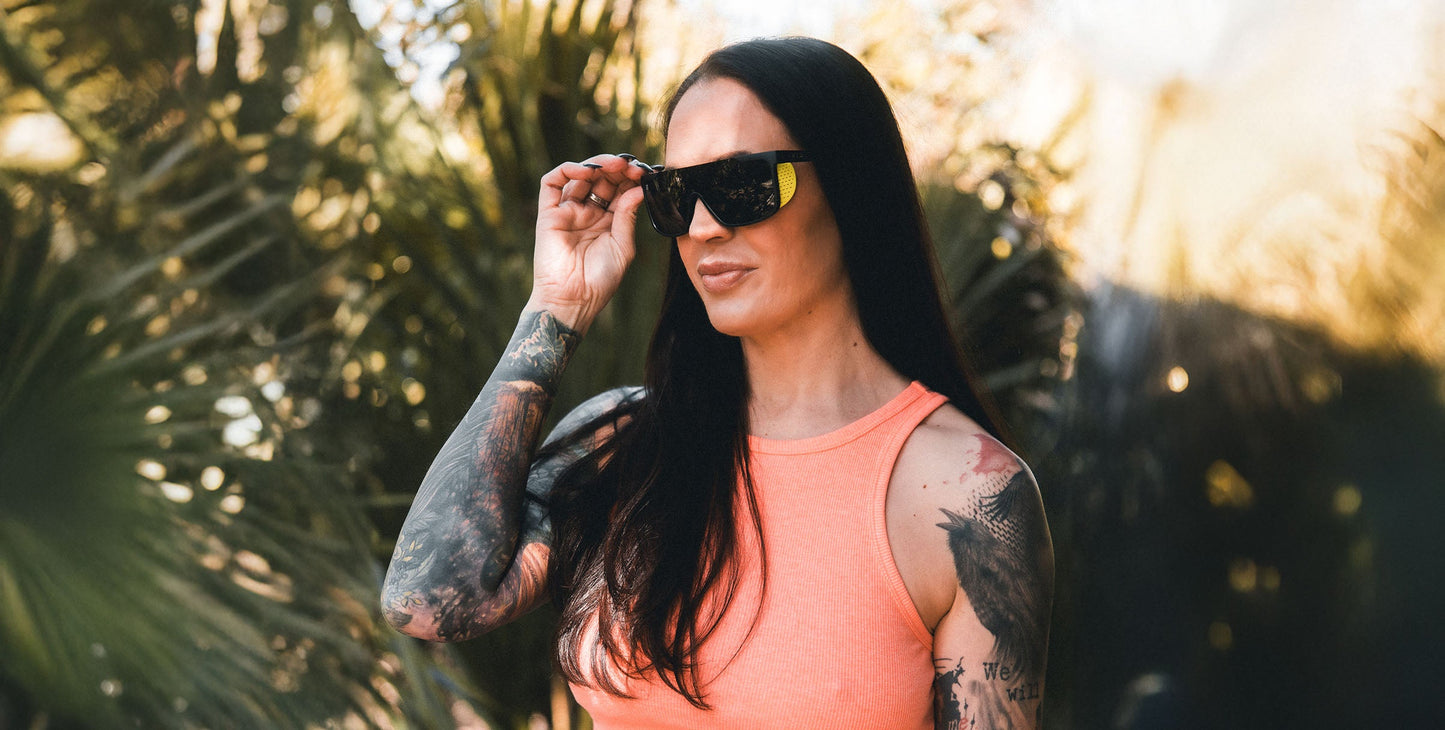 A woman with long dark hair and tattooed arms wears Neven Eyewear’s sleek Whistler sunglasses with polarized UV400 protection and a sleeveless coral top, smiling softly while standing outdoors among blurred green foliage and sunlight.