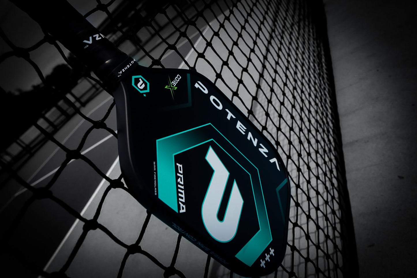 A close-up of the black and teal PRIMA COREx paddle by Potenza Pickleball rests against a chain-link fence on an outdoor court, highlighting its geometric design and bold logos.