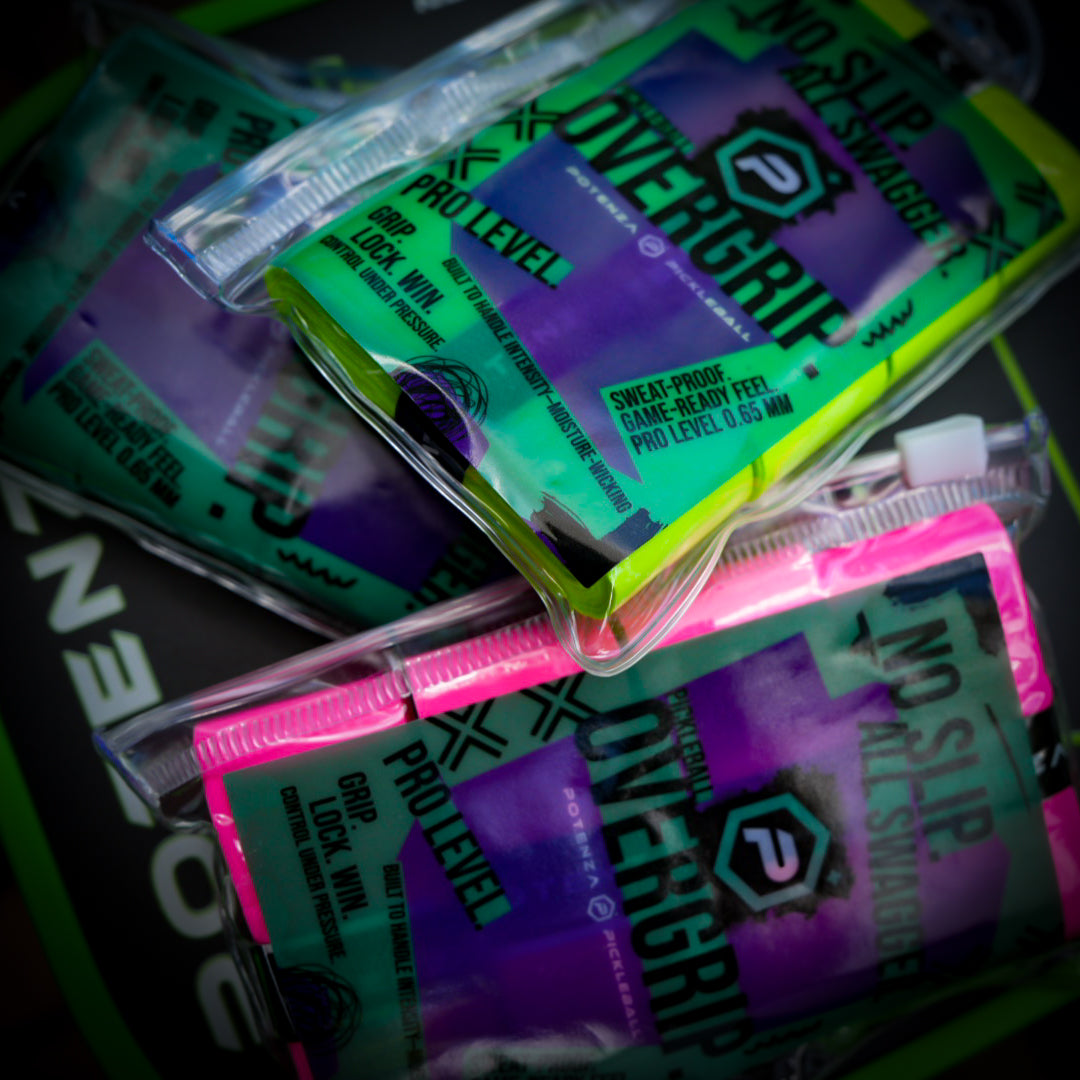 Close-up of colorful Potenza Pickleball PROX Tacky Overgrip (4-pack) packages—one green, one pink—with bold text and geometric designs, stacked at angles on a dark, patterned background.