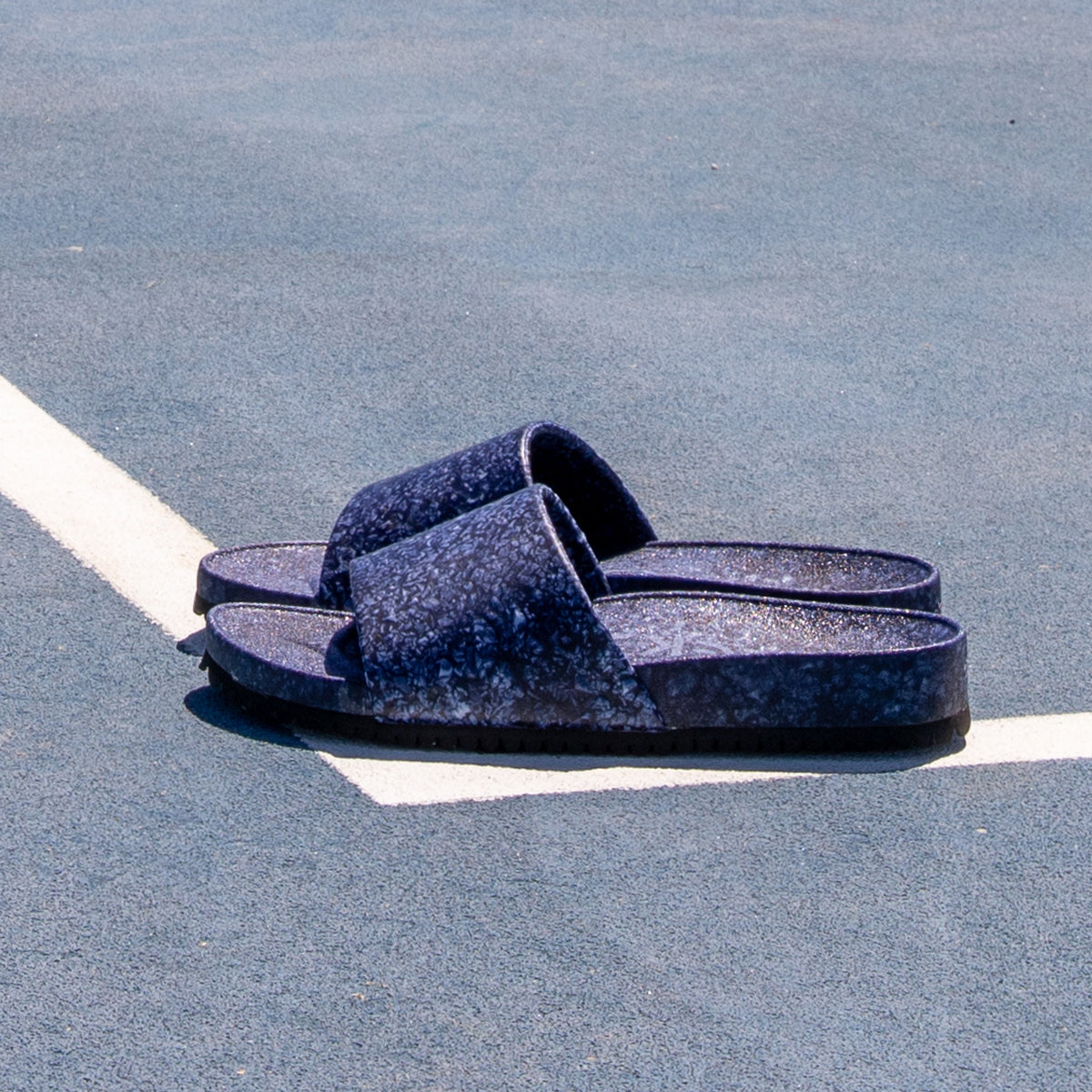 The Blumaka Fleks® East Beach Slide in dark blue, featuring a textured design for proper alignment, rests on a blue sport court at the intersection of two white lines, casting a shadow in bright sunlight.