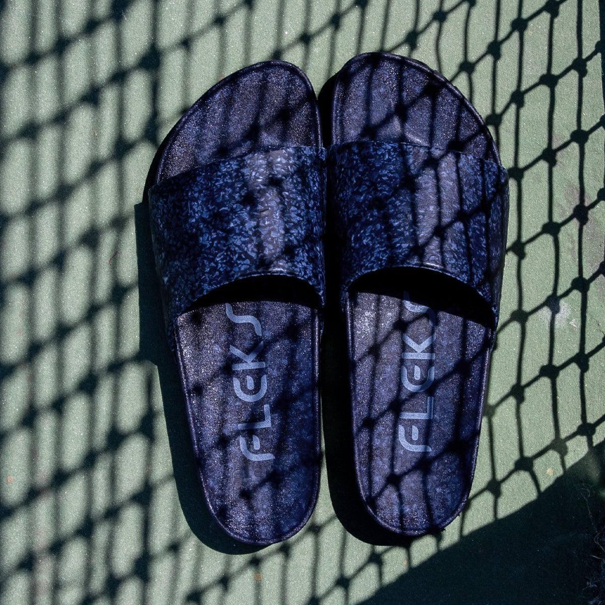A pair of Blumaka Fleks® East Beach Slide sandals in dark blue, featuring FLEKS on the footbeds, rests on a green surface beneath crisscross shadows from a chain-link fence. Designed for optimal alignment support.