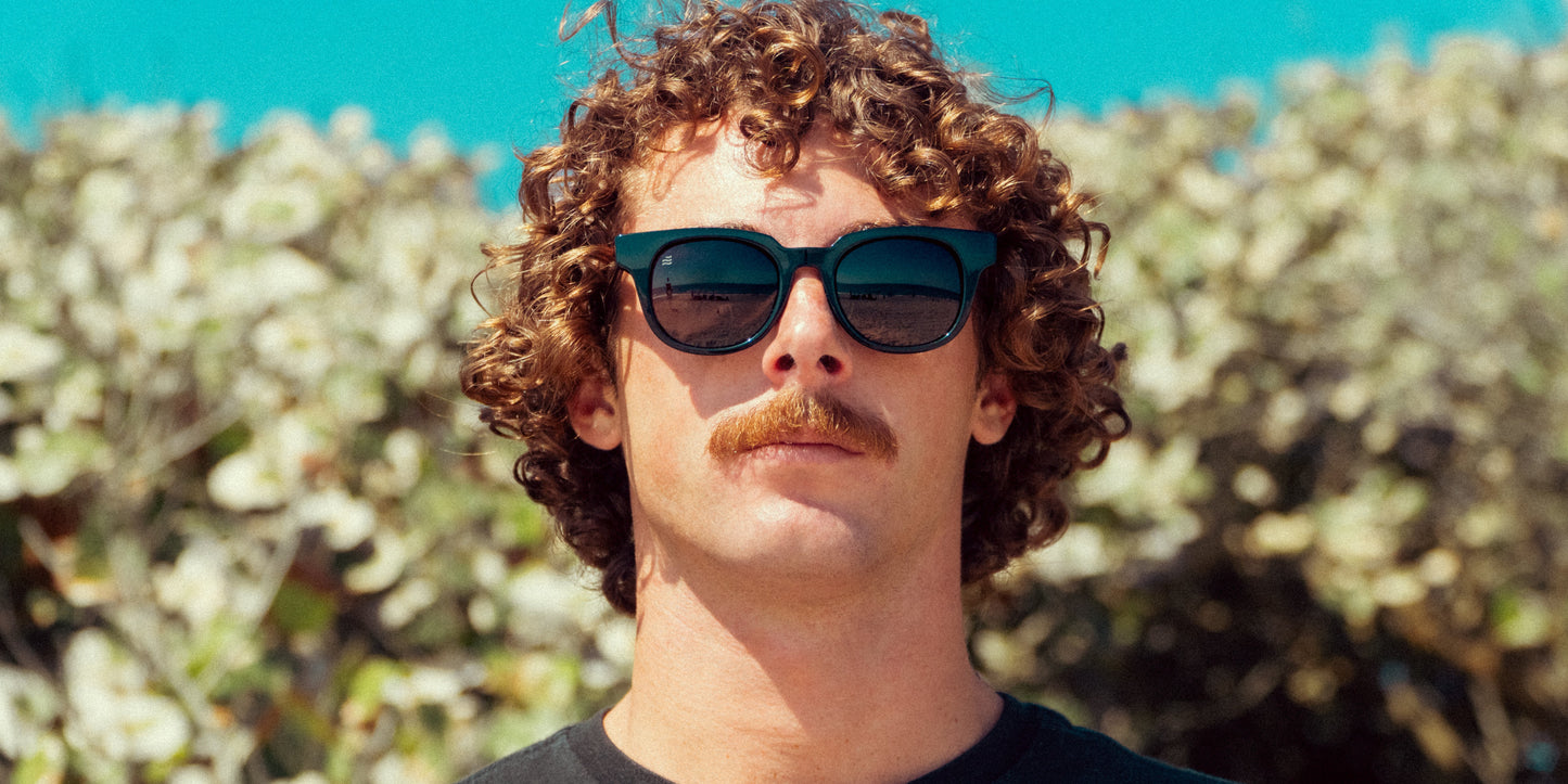 A man with curly brown hair and a thick mustache wears Neven Eyewear Cerberus sunglasses with black polarized lenses, reflecting a beach scene. He stands outdoors before sunlit green shrubs and a bright blue sky in a dark shirt.