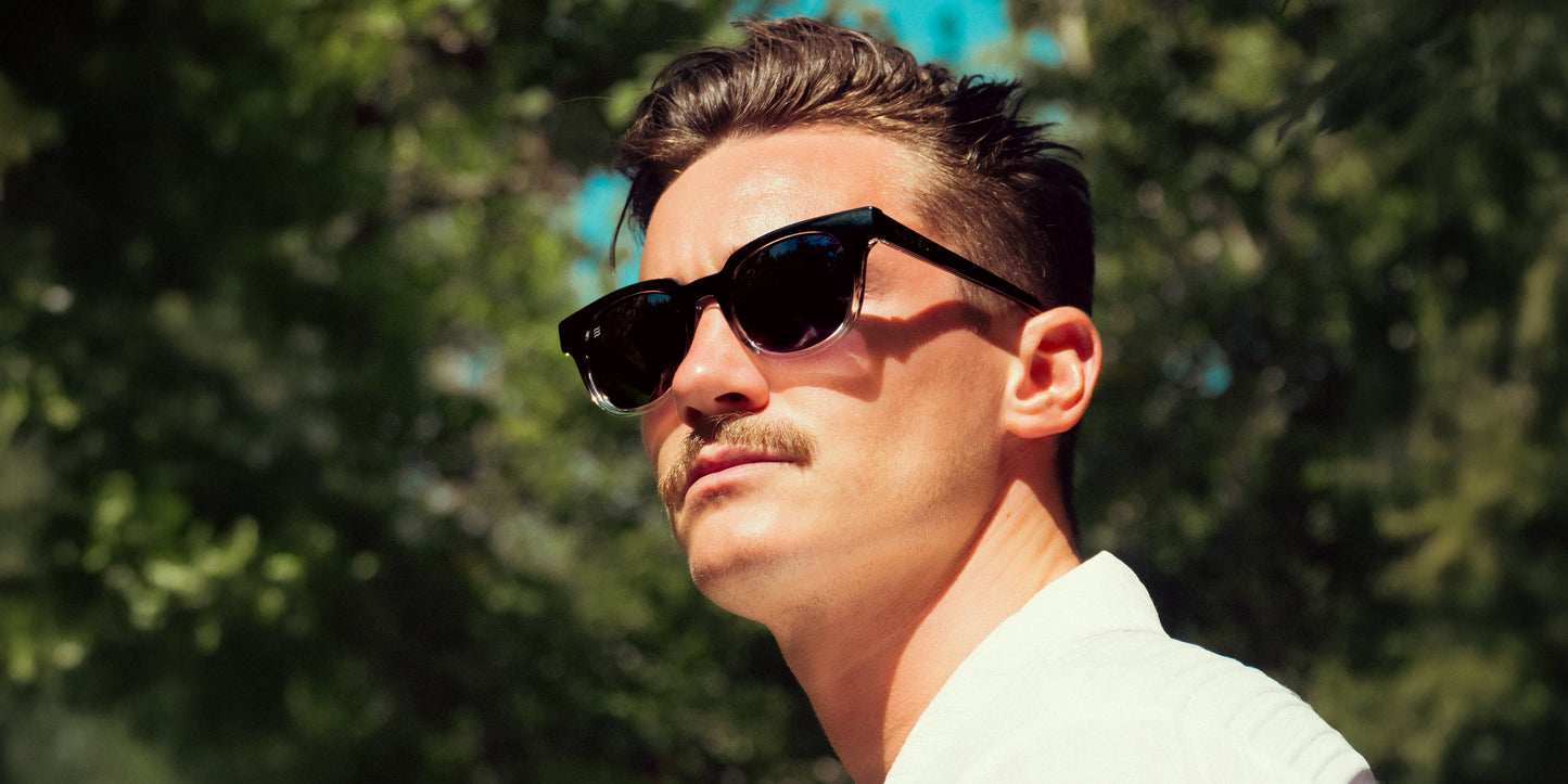 A man with short brown hair and a mustache stands outdoors in a white shirt, wearing Neven Eyewear’s Fang sunglasses with square gradient frames. Sunlight highlights his face amid blurred green trees and blue sky.