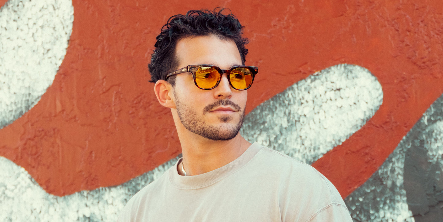 A man with short, dark curly hair and a light beard wears Neven Eyewear Gryphon sunglasses with yellow polarized lenses and a beige shirt, standing before a textured wall painted in abstract red, white, and gray patterns.
