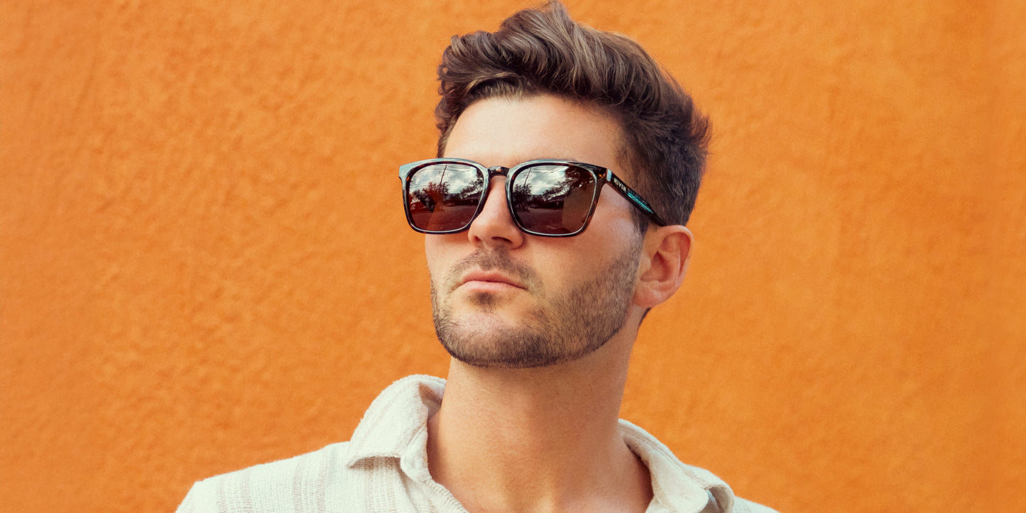A man with short brown hair and a trimmed beard wears Neven Eyewear’s Chazz tortoise shell sunglasses and a light shirt with an open collar, standing before a vibrant orange wall. Reflections of trees appear in his sunglasses.