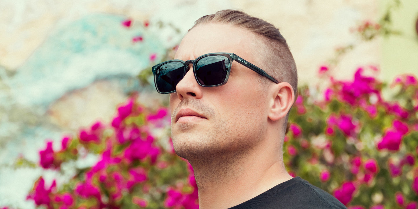 A man with light skin and short, slicked-back hair wears Neven Eyewear's Lenny black rectangular sunglasses and a black shirt, gazing upward amid bright pink flowers and blurred green foliage in sunlight.
