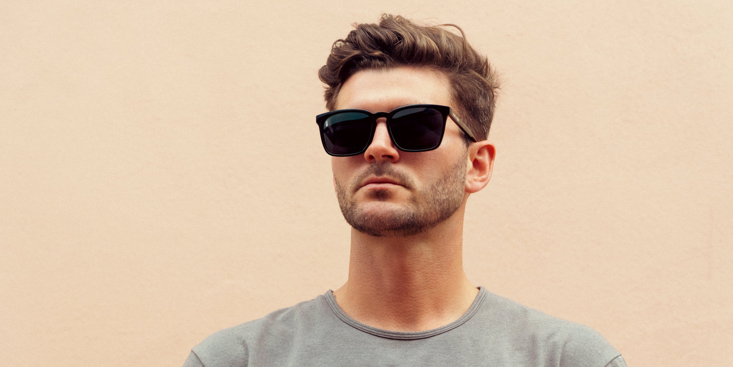 A man with short, wavy brown hair and a neatly trimmed beard wears Neven Eyewear’s Rufus polarized sunglasses and a light grey t-shirt. He stands against a pale peach background, gazing slightly to the side with a neutral expression.