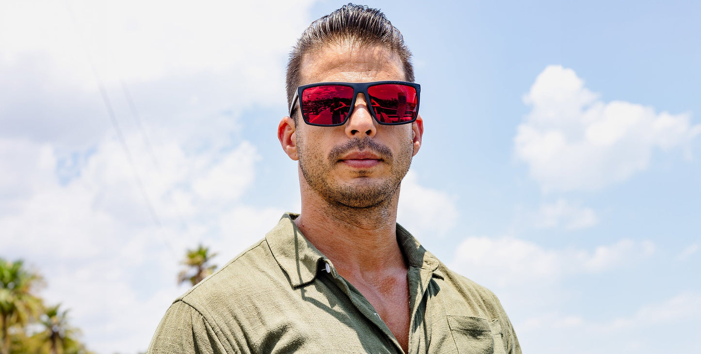 A man with slicked-back dark hair and stubble wears a green button-up shirt and the 212 sunglasses by Neven Eyewear. He stands outside on a sunny day, with blue sky, white clouds, and palm trees in the background.