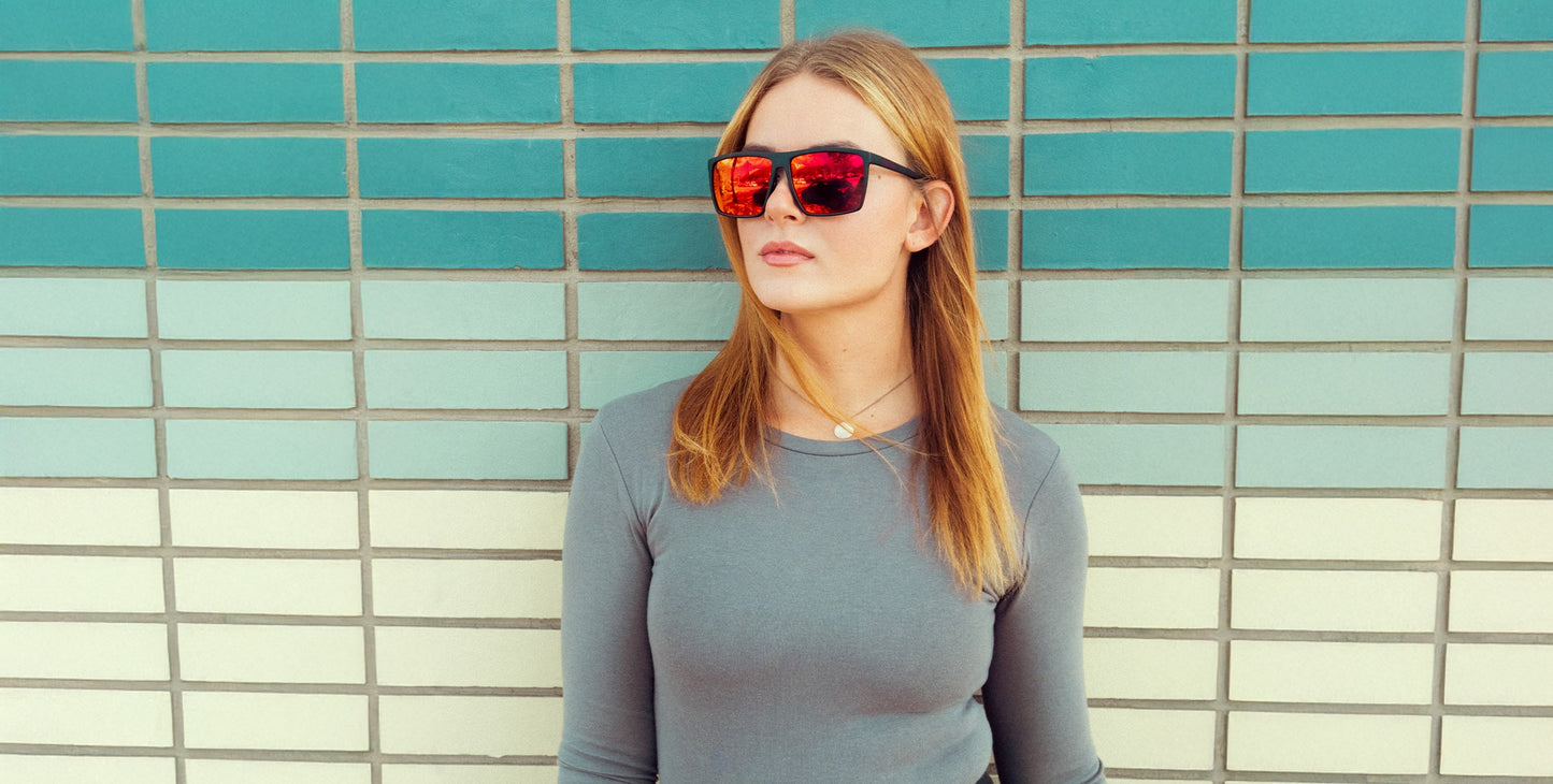 A young woman with straight, light brown hair wears Neven Eyewear's 212 red reflective glasses and a gray long-sleeve shirt, standing before a tiled wall of teal, turquoise, and light gray stripes while looking slightly aside.