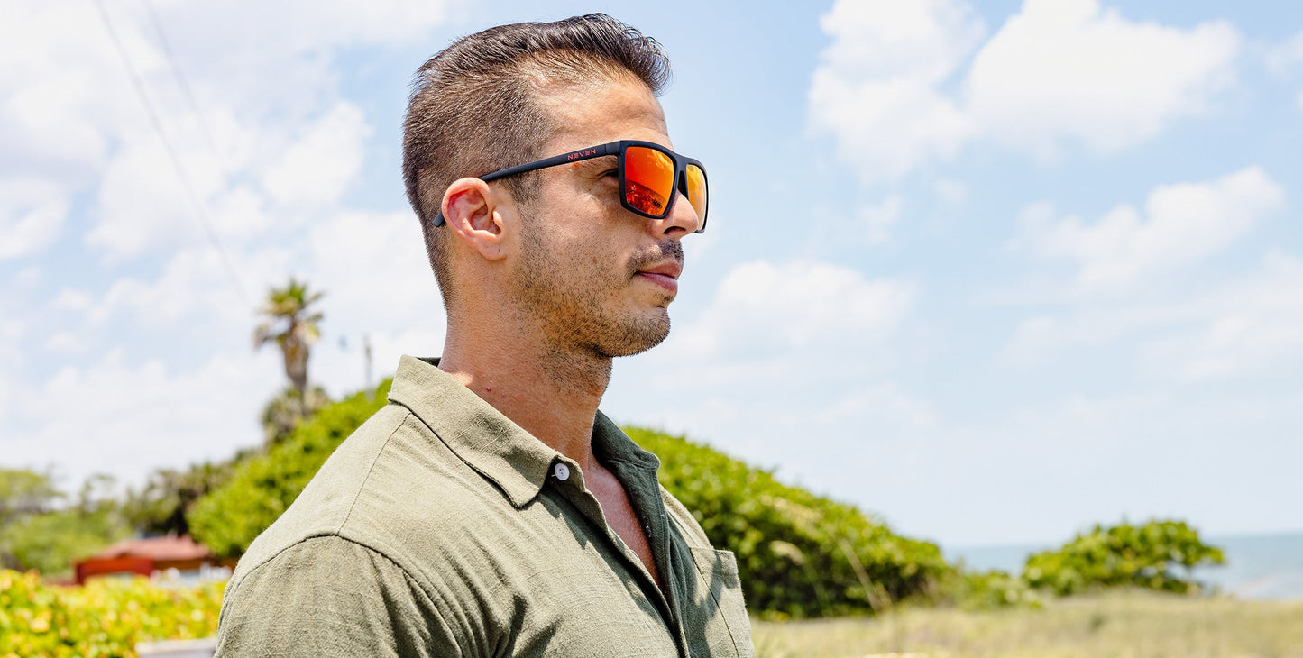 A man with short dark hair and stubble wears reflective 212 sunglasses by Neven Eyewear and a green collared shirt outdoors. Behind him are green bushes, a palm tree, partly cloudy sky, and distant ocean views.