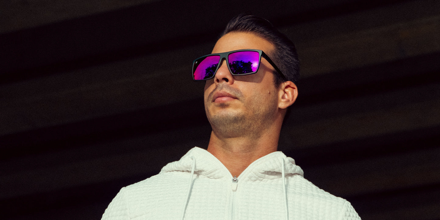 A man with short dark hair wears a textured white zip-up hoodie and Neven Eyewear 88th Street sunglasses featuring large, reflective purple polarized UV400 lenses with TR90 frames. He stands outdoors looking upward, with blurred dark beams in the background.