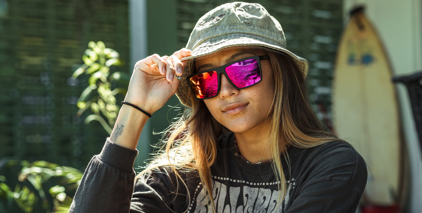 Outdoors amid greenery, a person sporting Neven Eyewear’s 88th Street sunglasses (polarized UV400, impact-resistant) and a green bucket hat adjusts their brim. A tattoo shows on their wrist; a surfboard lies nearby and they're wearing a dark graphic sweatshirt.