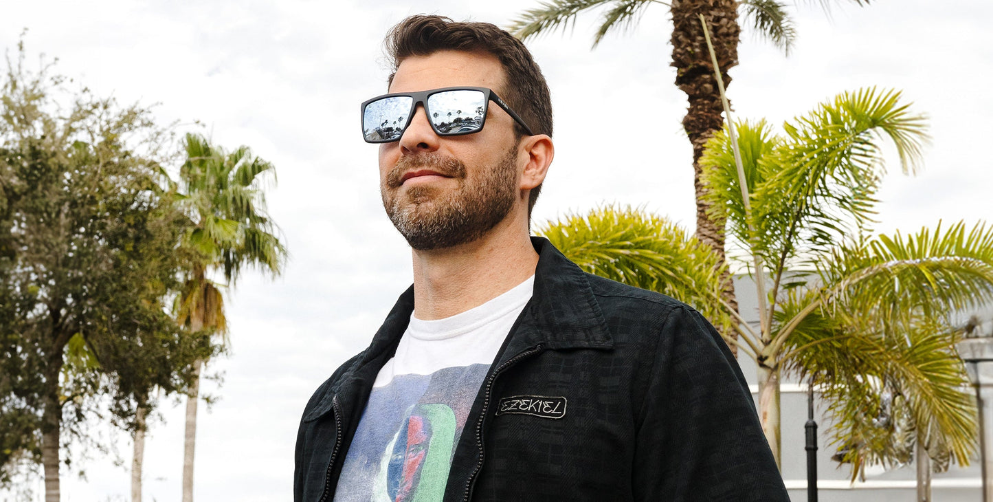 A bearded man wears Neven Eyewear’s August sunglasses with UV400 protection, plus a dark jacket and graphic tee. He stands among palm trees on a cloudy day, smiling slightly in the relaxed, overcast setting.