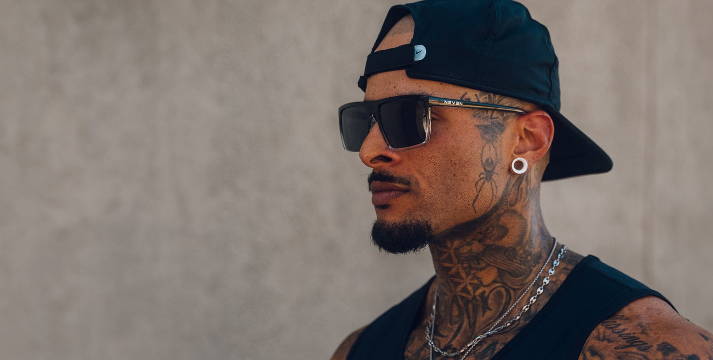 A tattooed man with a trimmed goatee wears Neven Eyewear's Colorado Bulldog sunglasses, featuring impact-resistant lenses, along with a black backwards cap and sleeveless shirt. He stands against a plain beige background, exuding a serious vibe.
