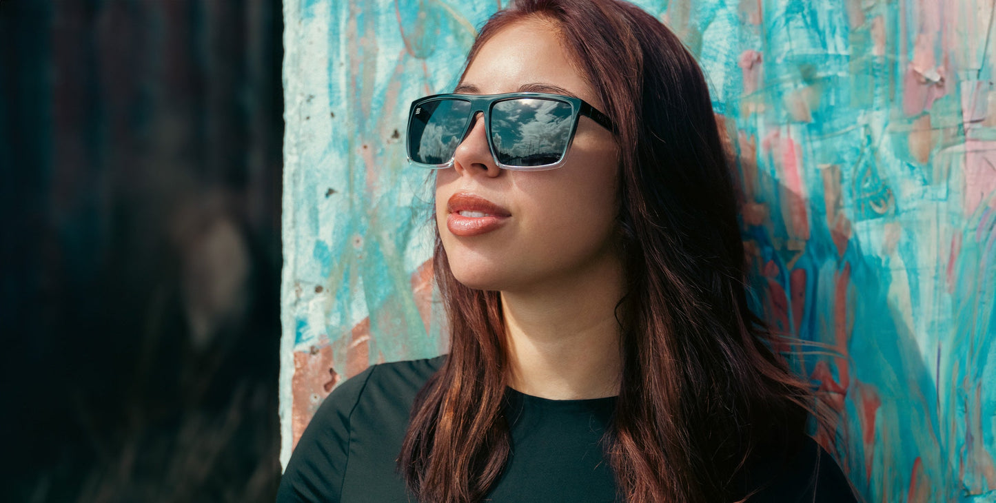 A confident woman in a black top and long brown hair leans against a graffiti wall, gazing upward. She wears Neven Eyewear's Colorado Bulldog sunglasses—large, polarized shades with UV400 protection that mirror the sky.