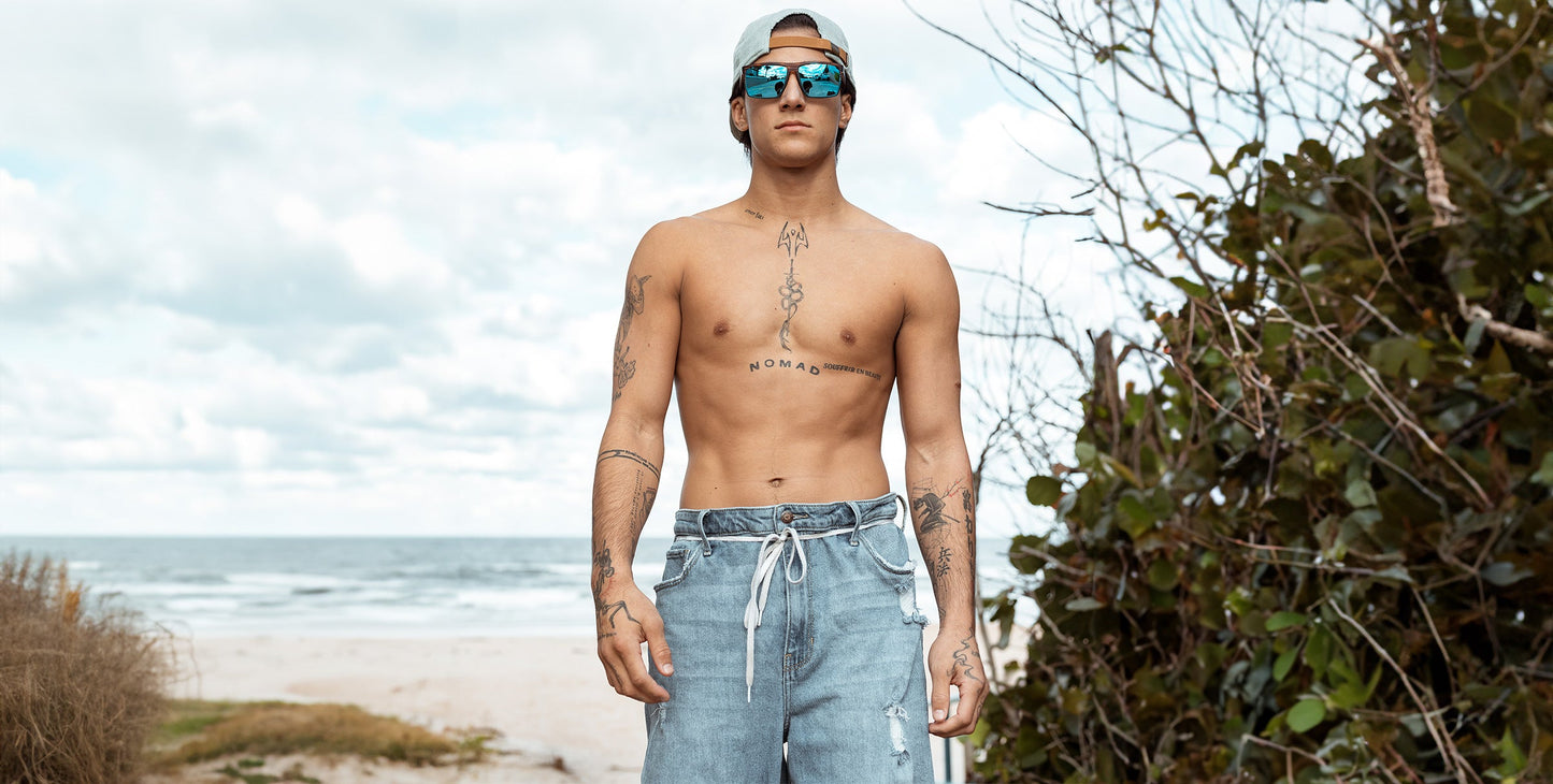 A shirtless man in blue denim shorts and a backwards cap stands on a sandy beach, wearing Neven Eyewear’s Melby polarized sunglasses with UV400 protection. He has multiple chest and arm tattoos. Green foliage and ocean waves are in the background.