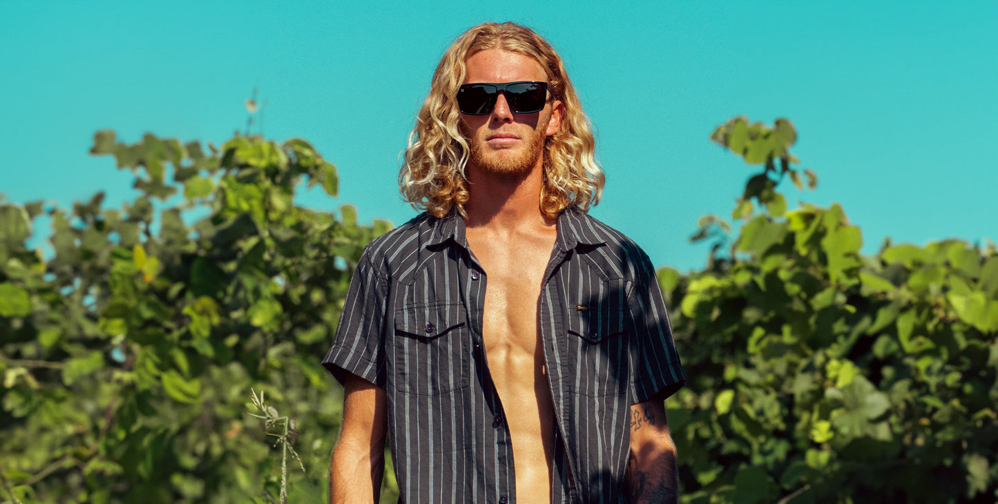 A man with long blond hair wears Neven Eyewear’s NYC sunglasses and a short-sleeved, dark striped button-up shirt open over his bare chest, standing outdoors against green foliage under a bright blue sky.