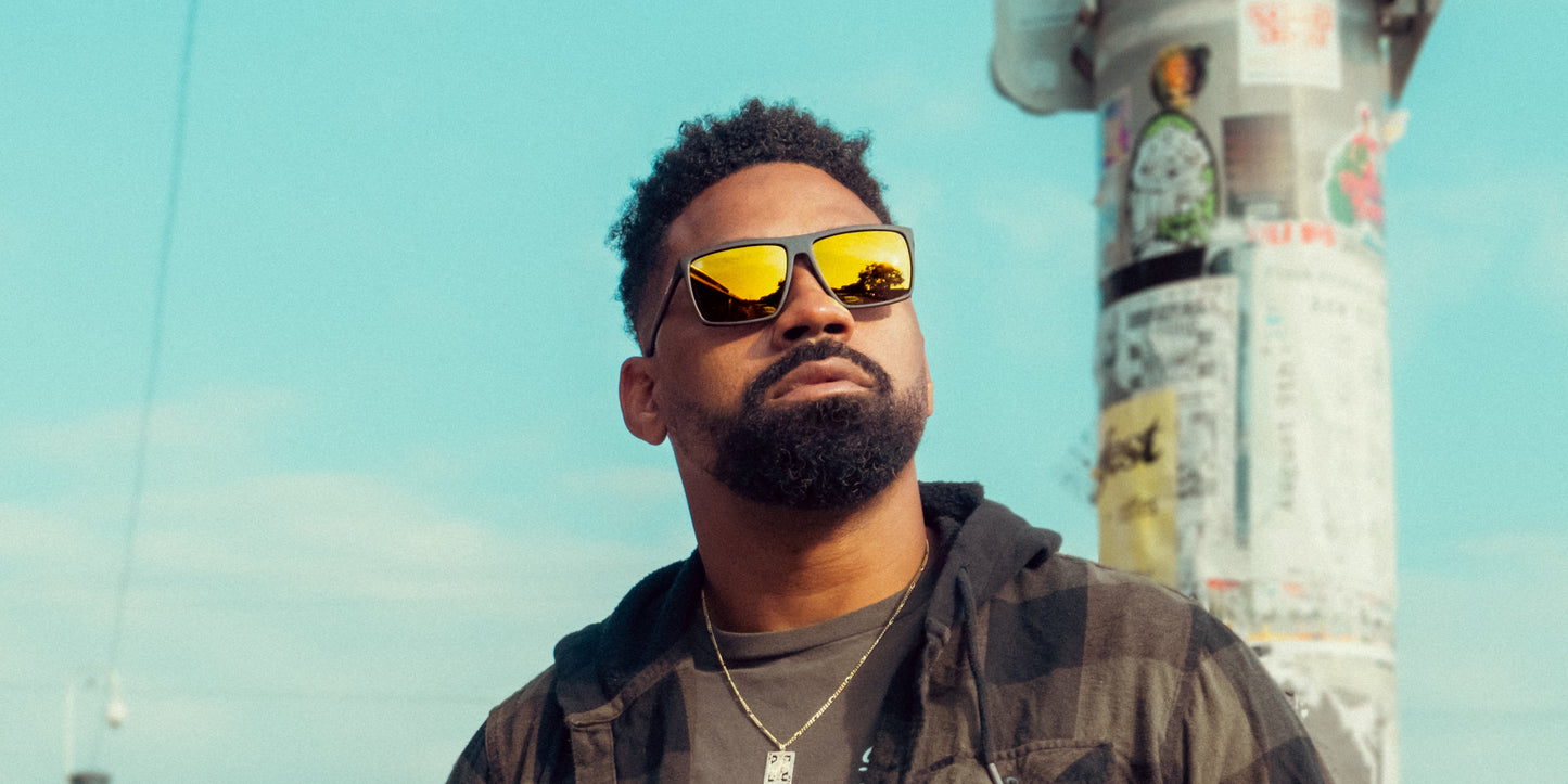 A bearded man in a plaid shirt wears Neven Eyewear’s Norton sunglasses. He gazes upward under a bright blue sky, with a sticker-covered pole behind him. The reflective lenses and vivid sunlight evoke a sunny day outdoors.