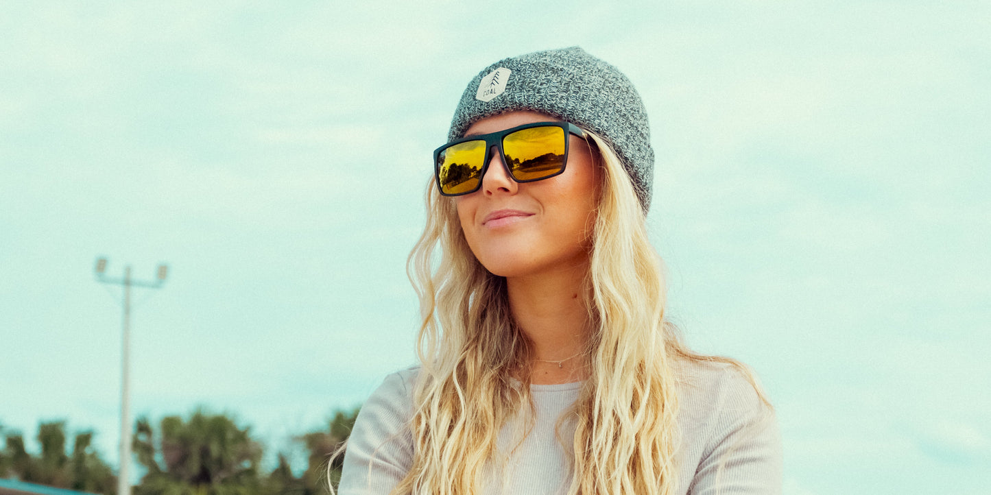 A person with long, wavy blonde hair wears a gray beanie and large mirrored Neven Eyewear Norton sunglasses. They’re dressed in a light gray shirt outdoors, with a clear sky, trees, and a light pole in the background.