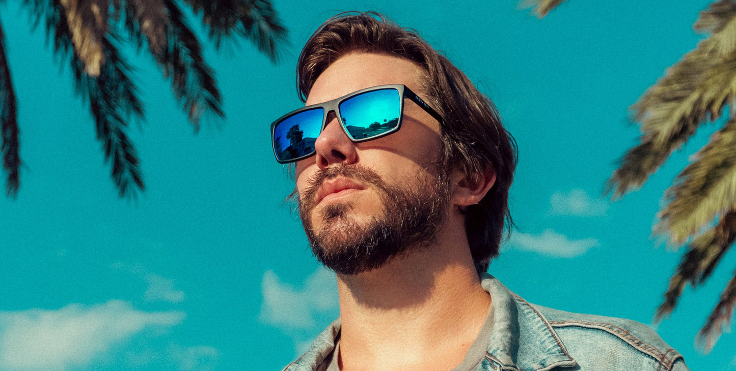 A bearded man in mirrored, polarized Three Of Cups sunglasses by Neven Eyewear and a denim jacket looks up, palm trees and blue sky reflected in his UV400 lenses, capturing a bright, summery vibe.