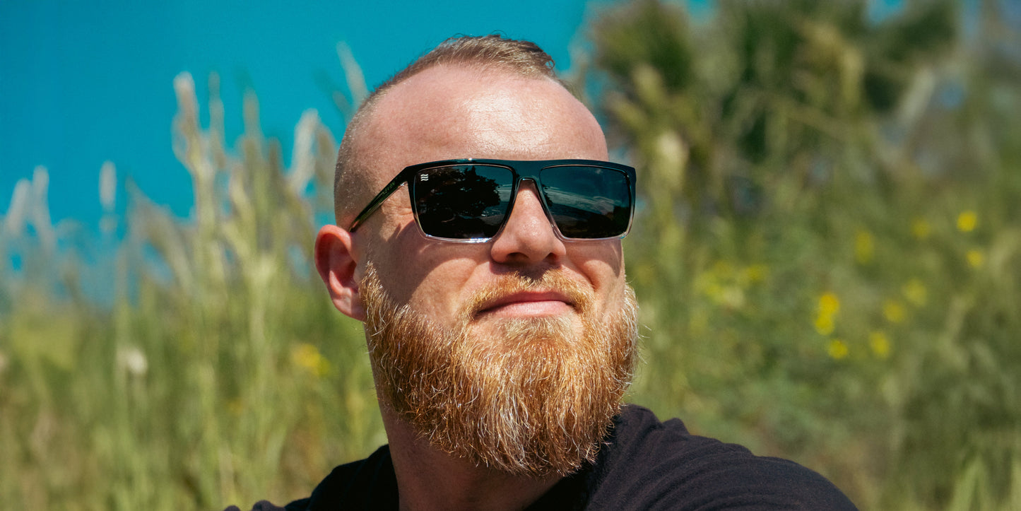 A bearded man with short hair wears Neven Eyewear’s Colorado Bulldog XL prescription sunglasses and a black shirt, relaxing outdoors in the sunlight. Tall grasses, green foliage, and a blue sky form the backdrop.