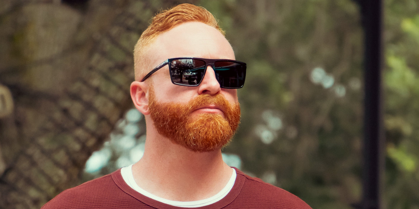 A bearded man with neat red hair wears Neven Eyewear's NYC XL black rectangular sunglasses and a maroon sweater over a white shirt, standing outdoors against a blurred leafy background with a metal fence partly visible.
