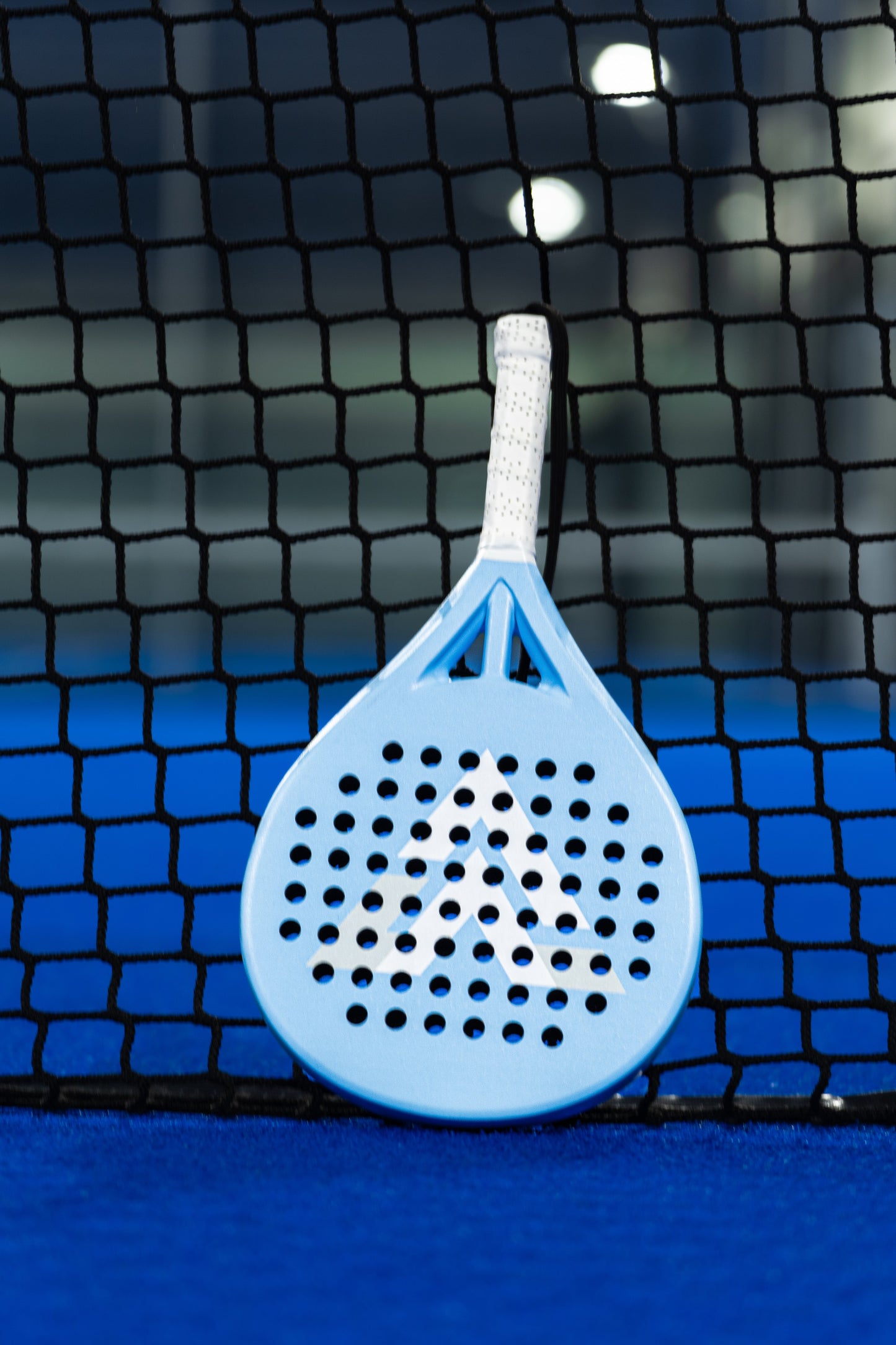 The VALA One (Junior Edition) padel racket by VALA, featuring a light blue frame, white grip, and arrow pattern, stands upright against a black net on a bright blue padel court—ideal for young players.