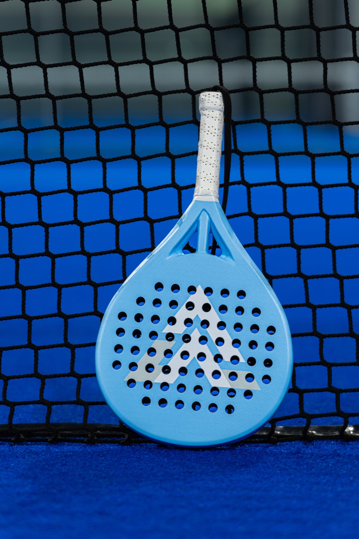 The VALA One (Junior Edition) by VALA, featuring a blue frame with a white geometric design and perforated holes, leans against a black net on a bright blue court—an ideal starter padel racquet for kids.