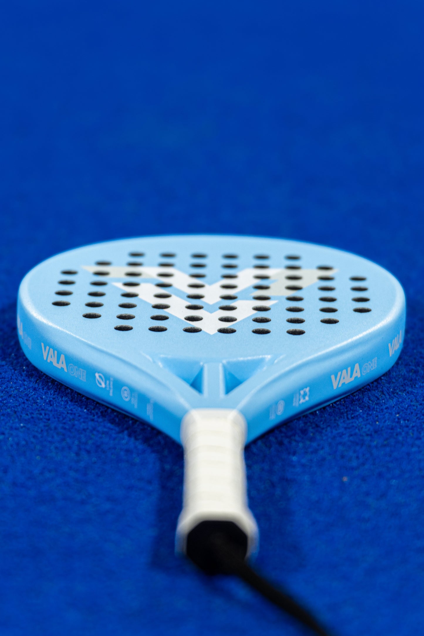 A close-up of the VALA One (Junior Edition) padel racket by VALA, with a white grip, black perforated holes, and a white W design, lies on a bright blue court. The handle is slightly blurred in the foreground.