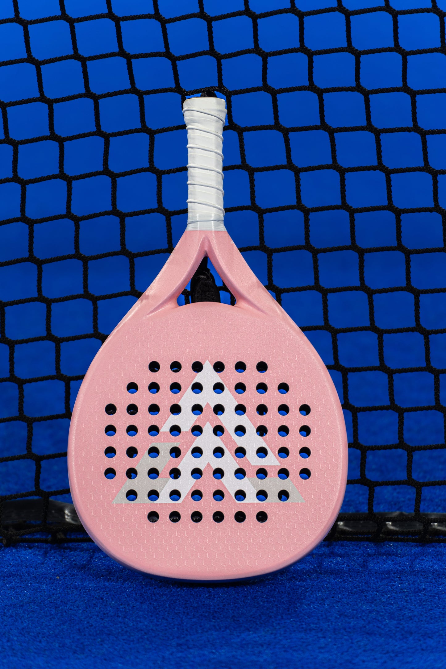 The VALA One (Pink Edition) by VALA is a premium light pink padel racquet with a white grip and EVA foam core, standing upright against a black net on a blue court. A white geometric triangle logo features at the center.
