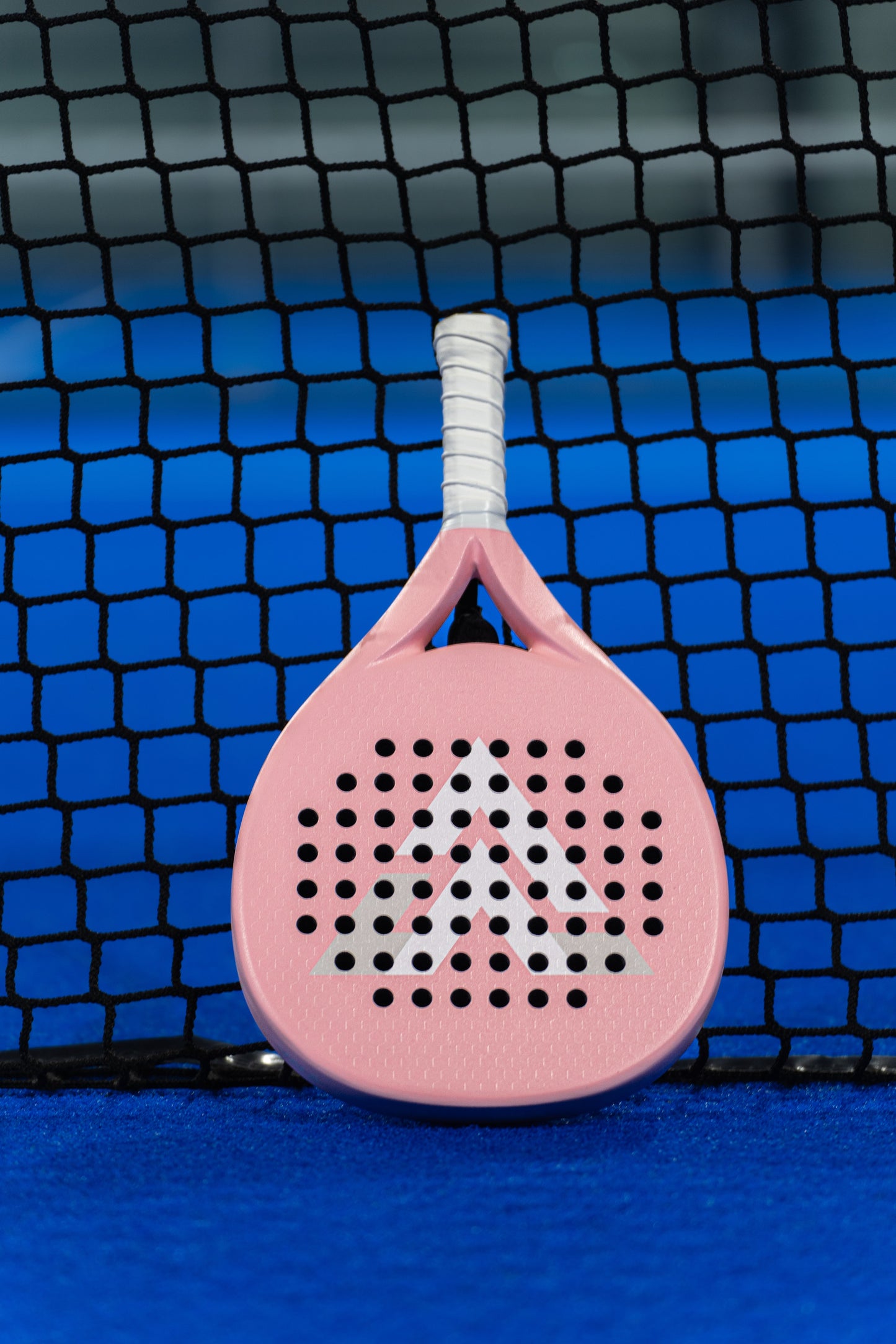 The VALA One (Pink Edition) padel racquet by VALA features a light pink design, white handle, chevron accents, an EVA foam core, and rows of holes across the face. It is shown upright against a black net on a vibrant blue court.