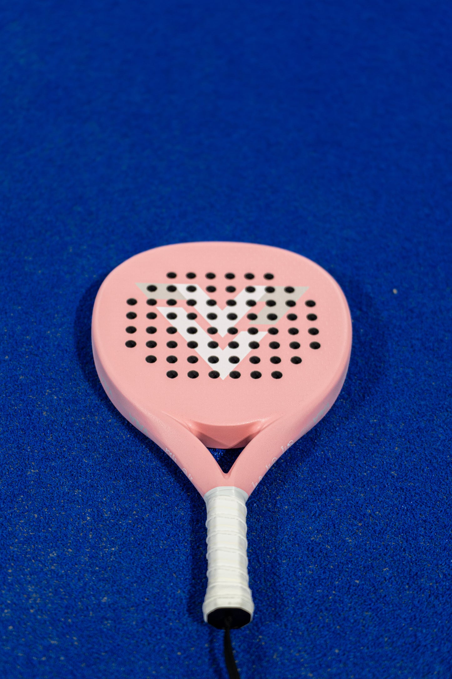 The VALA One (Pink Edition) padel racquet by VALA features a light pink design with a white grip, EVA foam core, round holes, and a bold white V. It rests on a bright blue court, its vibrant colors and clean lines highlighted in the light.