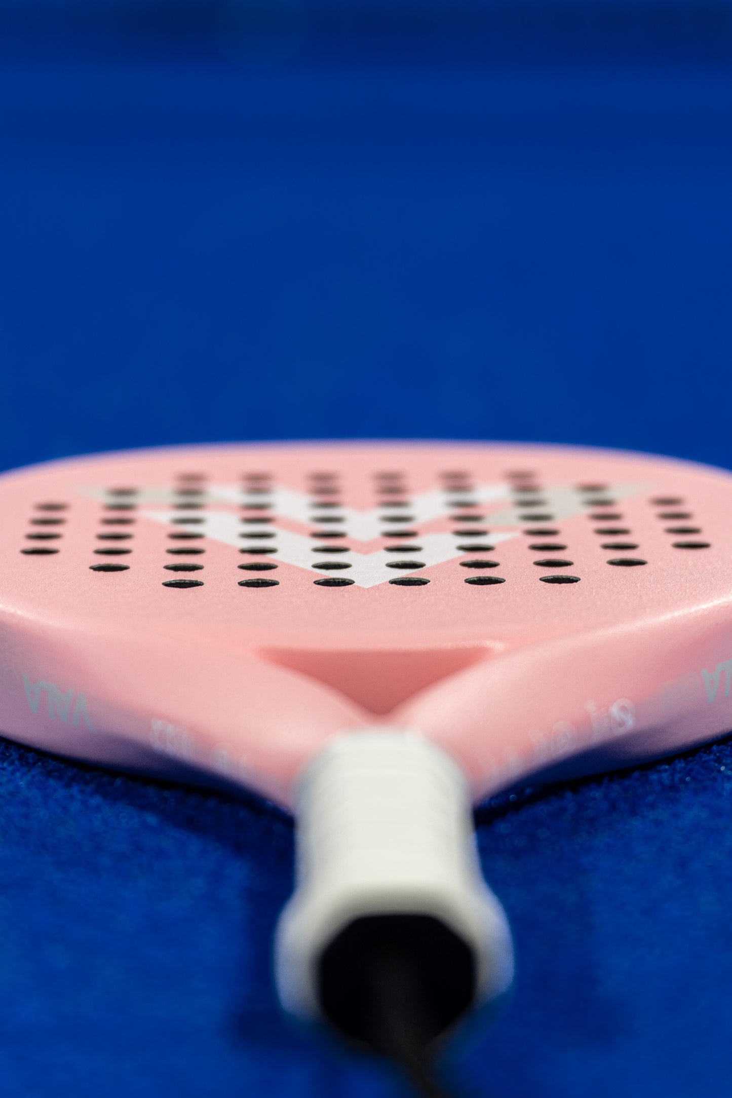 Close-up of the VALA One (Pink Edition) padel racquet by VALA, in light pink with black holes and a white handle, featuring an EVA foam core. The racquet lies flat on a bright blue court, highlighting its premium design.