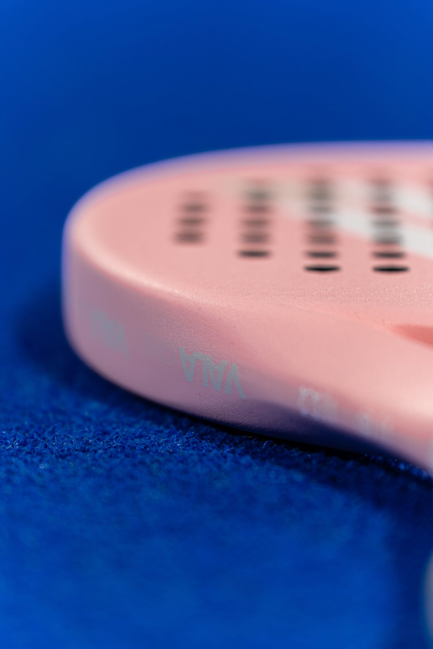 A close-up of the VALA One (Pink Edition) padel racquet by VALA, featuring an EVA foam core, rests on a textured blue court. Visible small holes and subtle branding highlight its premium design and bold pink color.