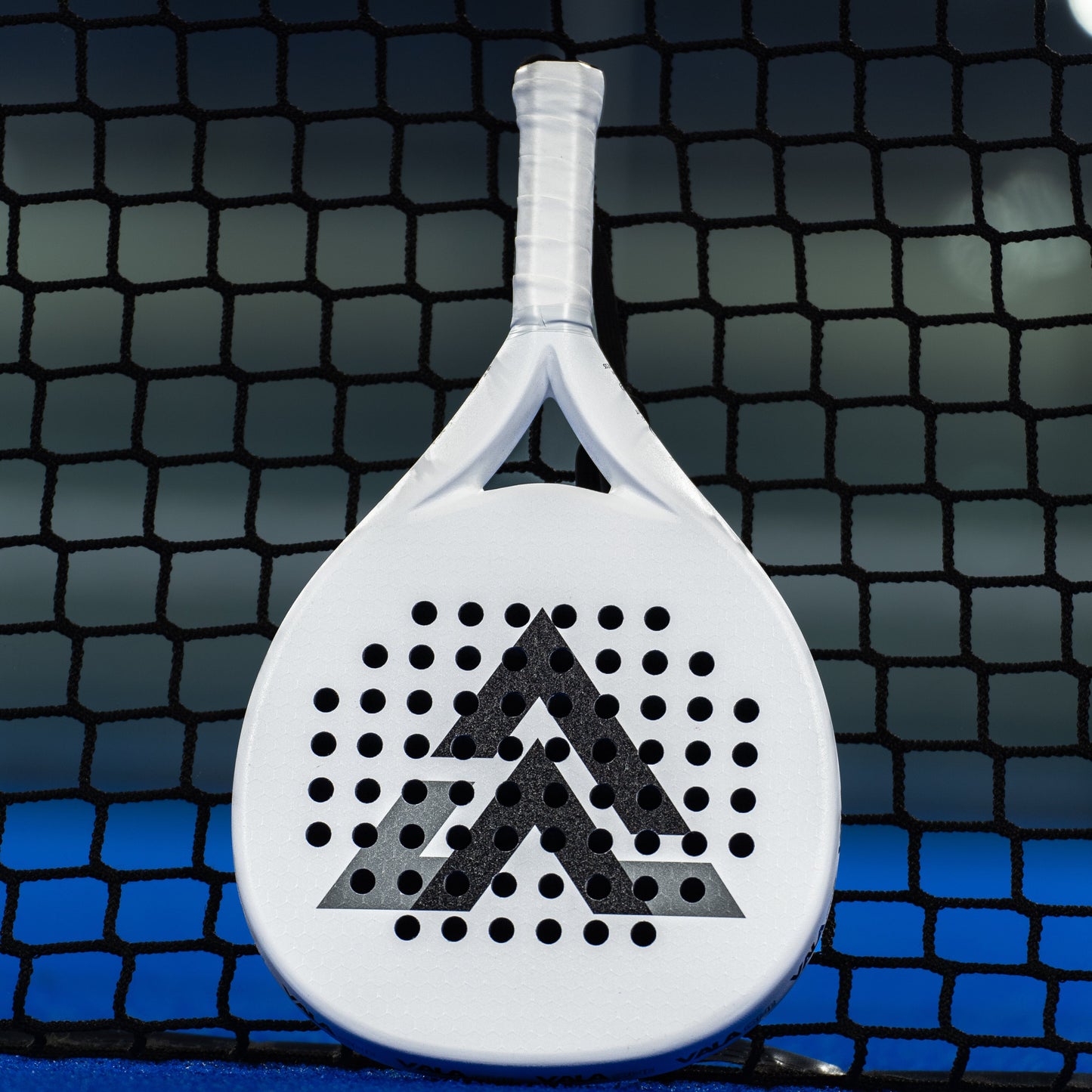 The VALA One (White Edition) padel racquet by VALA stands upright on a blue court, featuring a minimalist geometric black arrow design and circular holes, highlighted against a black net and blurred background.