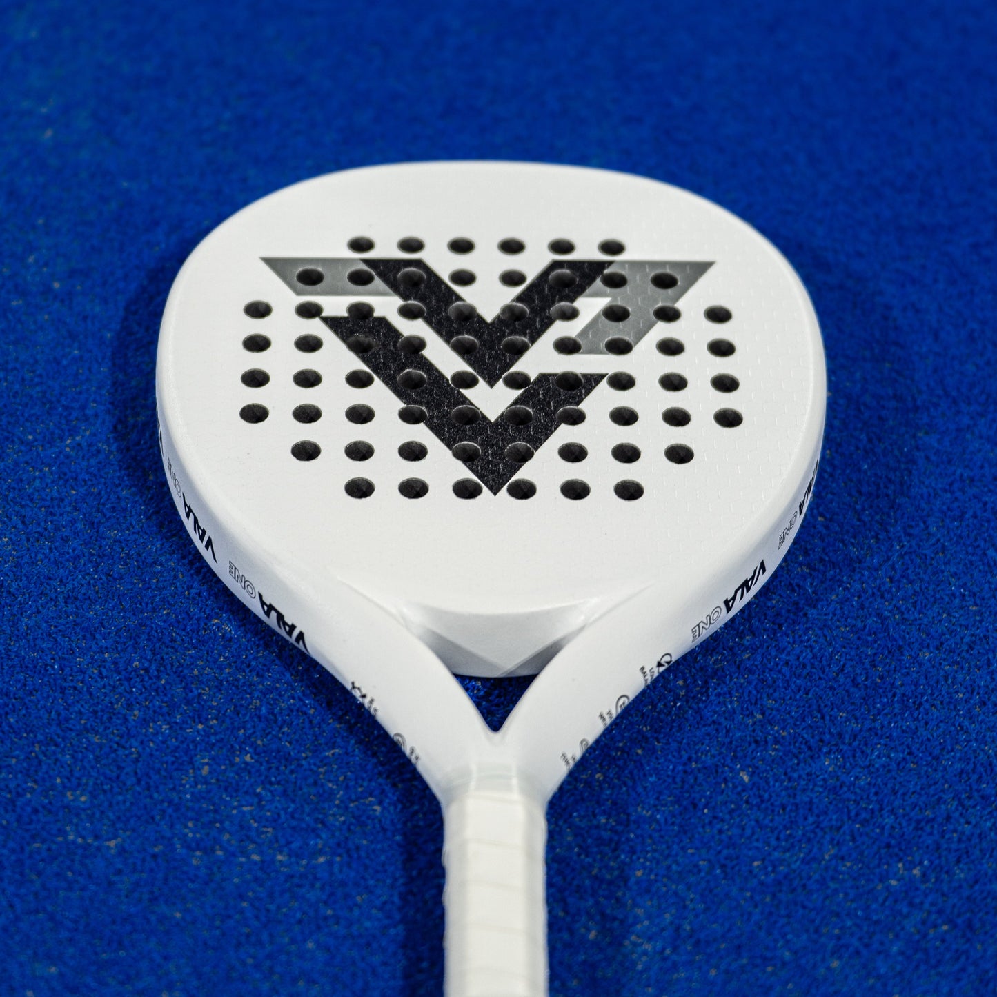 The VALA One (White Edition) padel racquet by VALA features a minimalist white design with a bold black “V” logo and circular holes, complemented by a wrapped handle. It rests elegantly on the bright blue textured court.