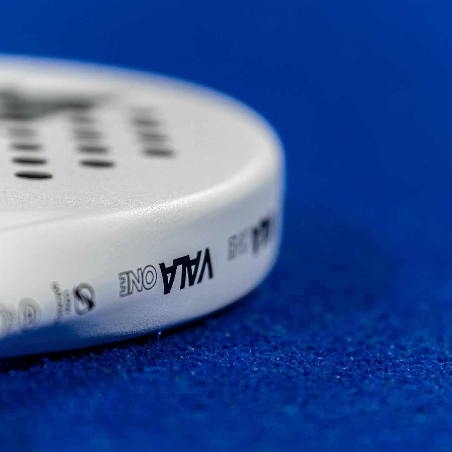 Close-up of the VALA One (White Edition) padel racquet by VALA, featuring small top holes and sleek branding, shown resting on a textured blue surface that highlights its minimalist design.