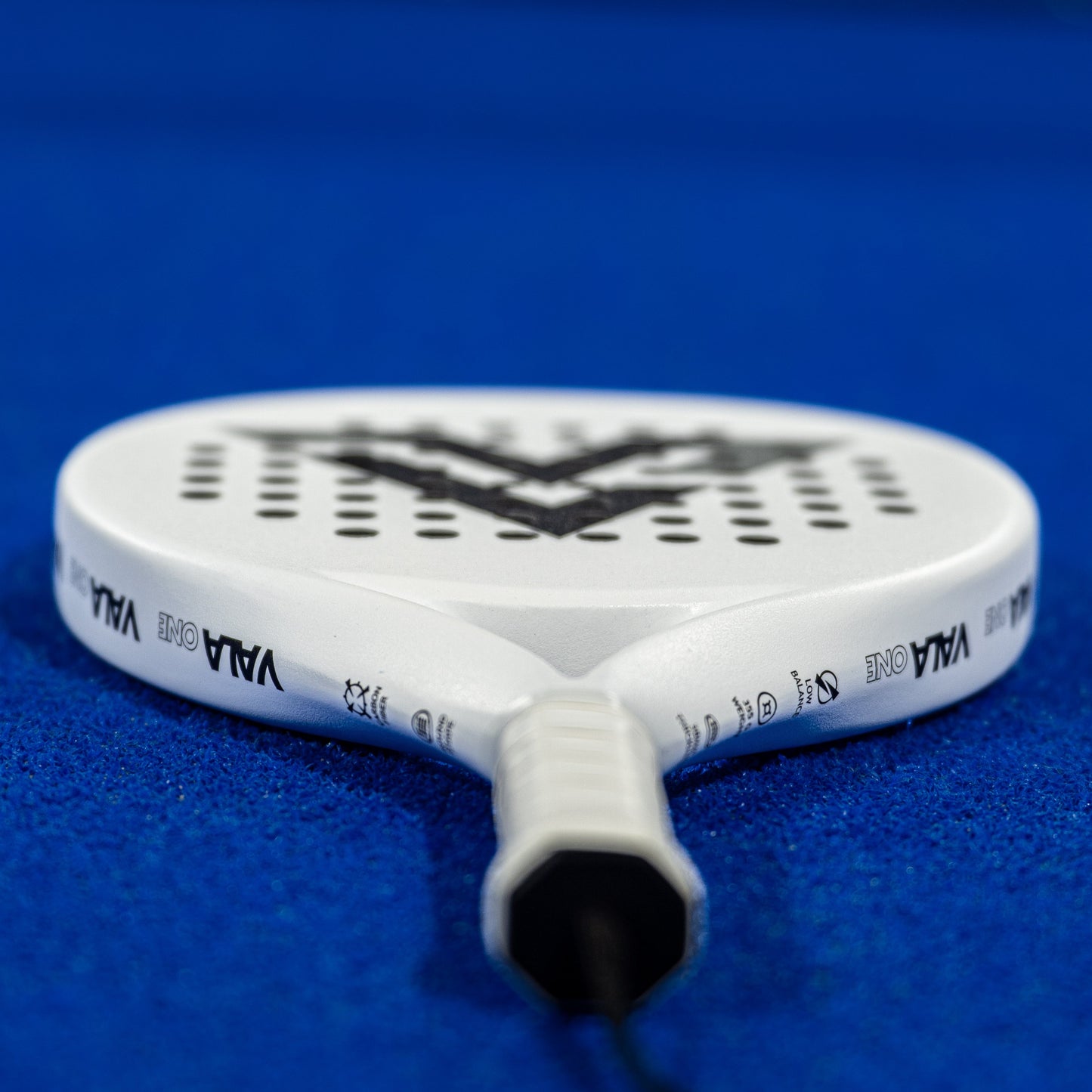 A close-up of the VALA One (White Edition) padel racquet by VALA rests on a bright blue court, its handle facing the camera. The rounded face features bold VALA branding, complemented by black accents and perforations.