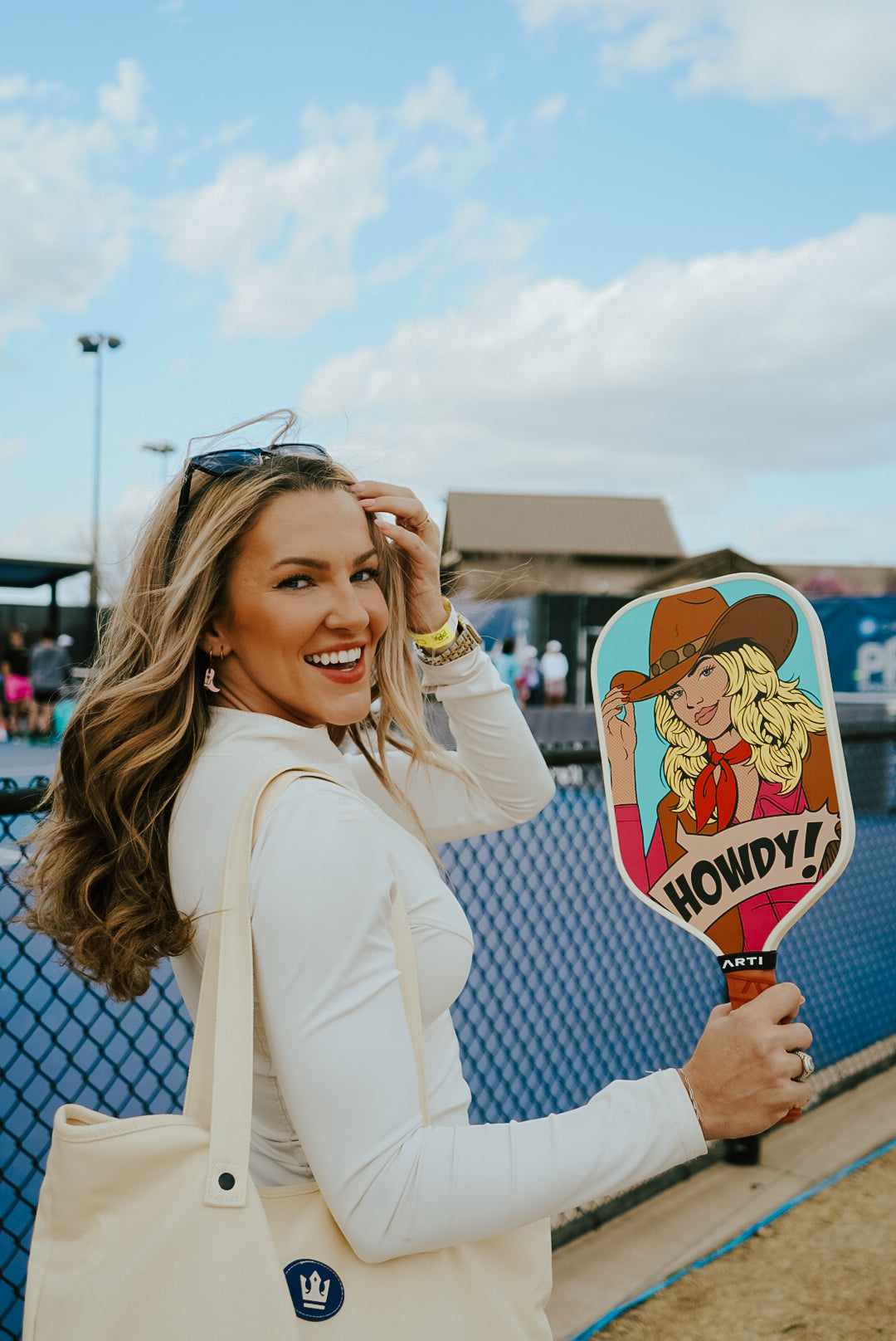 A smiling woman in white holds an ARTI Carbon Fiber Kristen & Kristy "HOWDY" Paddle by ARTI, featuring a cartoon cowgirl, and a tote bag. She stands outside near a blue chain-link fence, with people and buildings in the background under a blue sky.