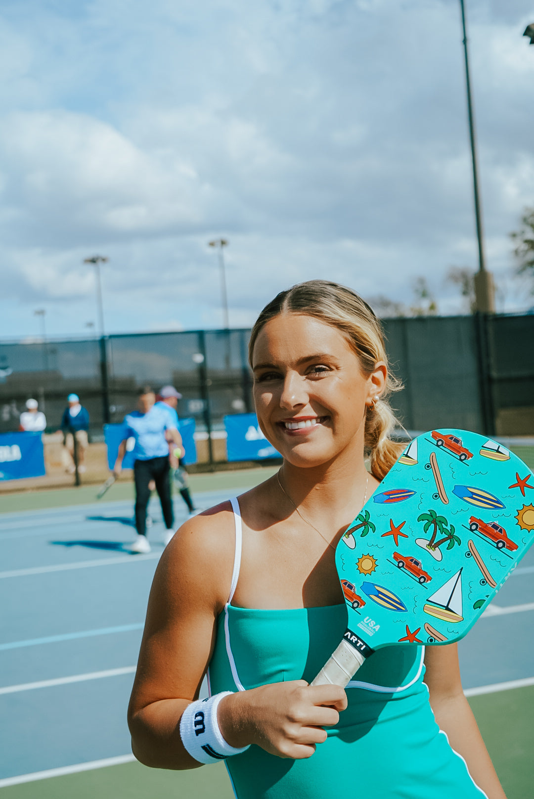 A young woman in a teal tank top smiles on an outdoor pickleball court, holding the ARTI Carbon Fiber "CALIFORNIA" Paddle by ARTI, featuring beach-themed art. She wears a white wristband; other players appear blurred in the background.