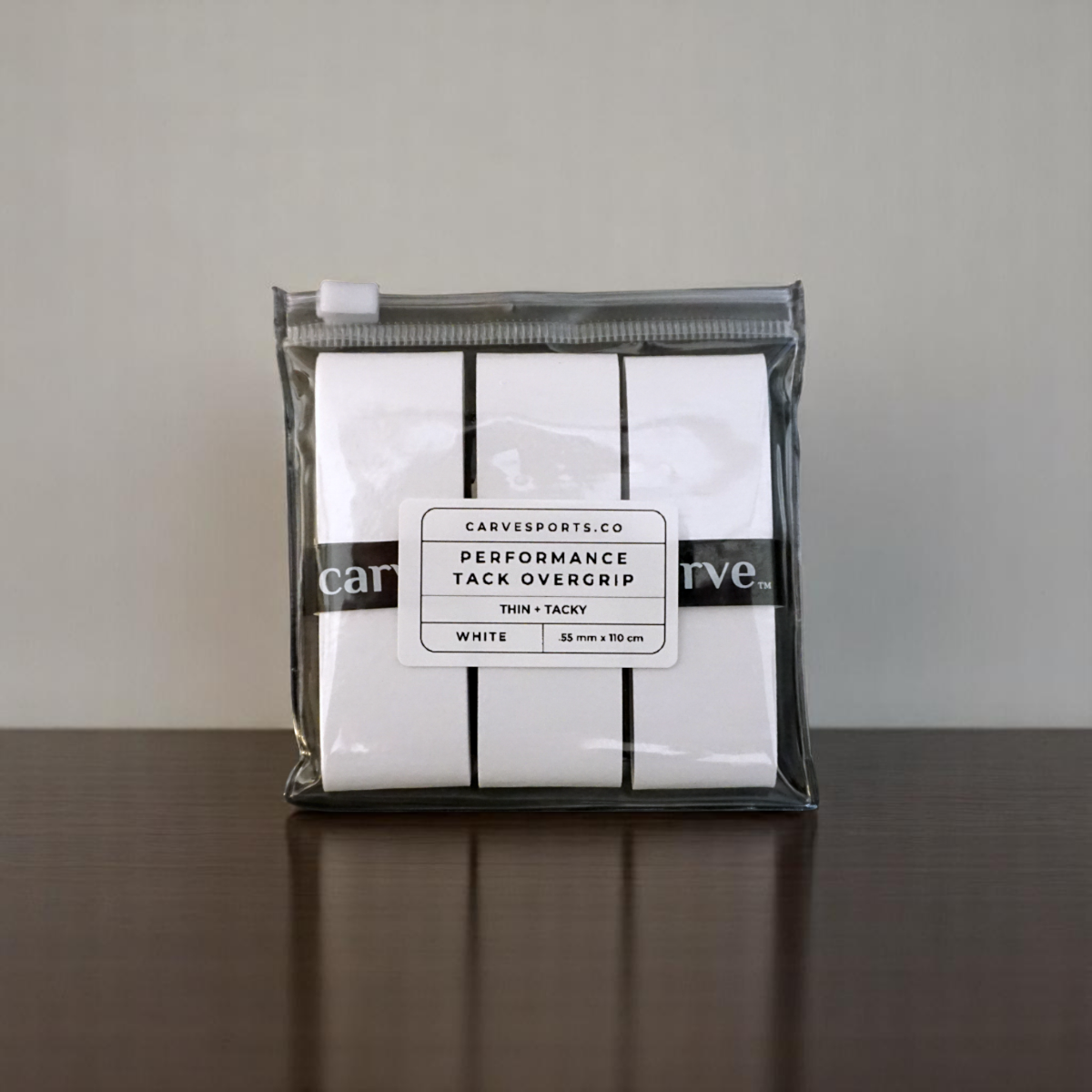 A clear pouch containing three white Carve Sports - Performance Overgrips for racket grip sits on a dark wooden surface. The label reads: “Carve Sports PERFORMANCE OVERGRIP, Thin-Tacky, 35mm x 110cm, White.” Pouch is sealed and neatly packaged.