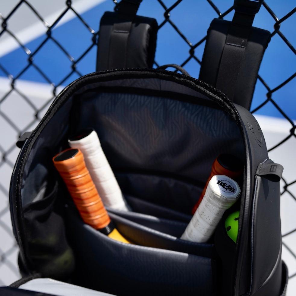 A close-up of the FORWRD Court Caddy Pickleball Bag, made from premium materials, hangs on a chain-link fence by a blue tennis court. Inside are three racquet handles—one orange, two white—and a bright green tennis ball.