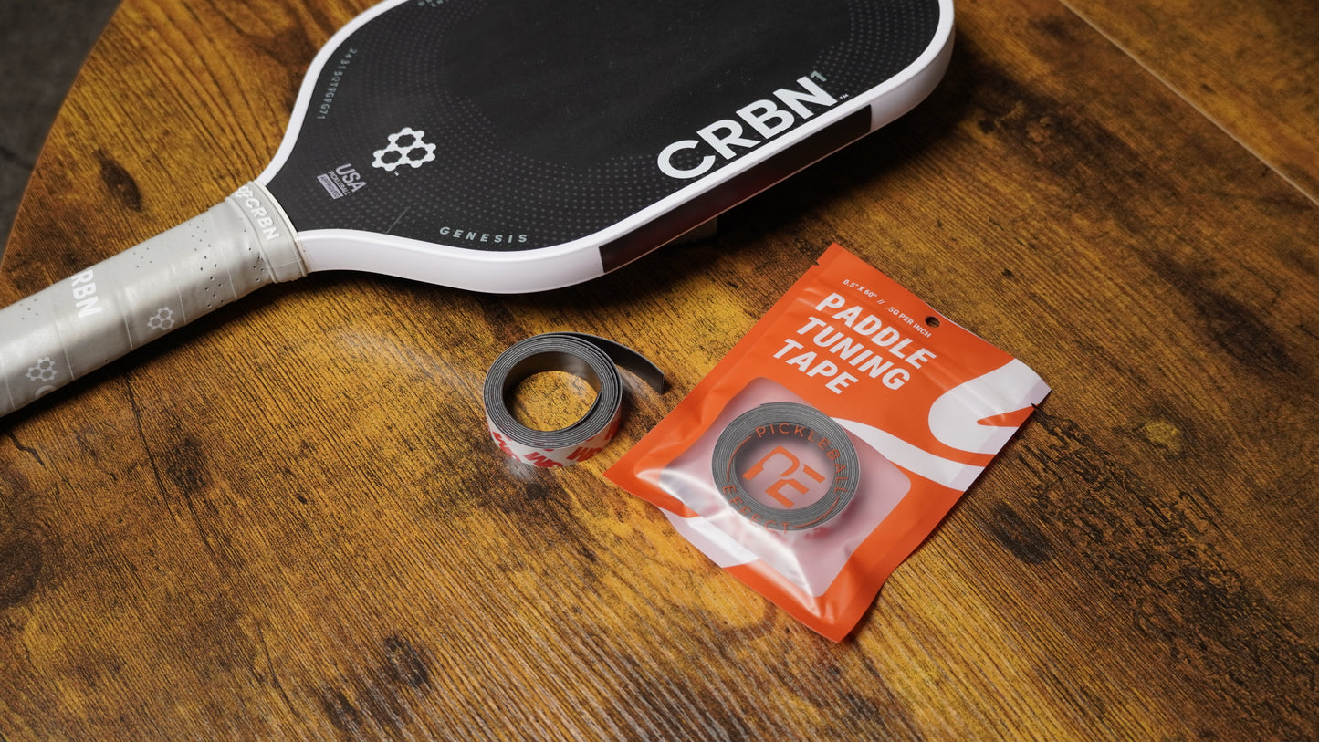 A CRBN pickleball paddle lies on a wooden surface next to two rolls of Pickleball Effect Shop Paddle Tuning Tape, one in its orange and white packaging, suggesting equipment maintenance or paddle weight adjustment.