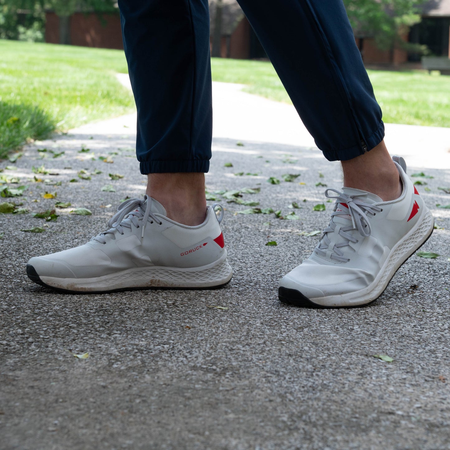 A person wearing dark blue pants and light gray athletic shoes with Blumaka Recovery Bundle walks on a paved path outdoors, with green grass, trees, and brick buildings blurred in the background.