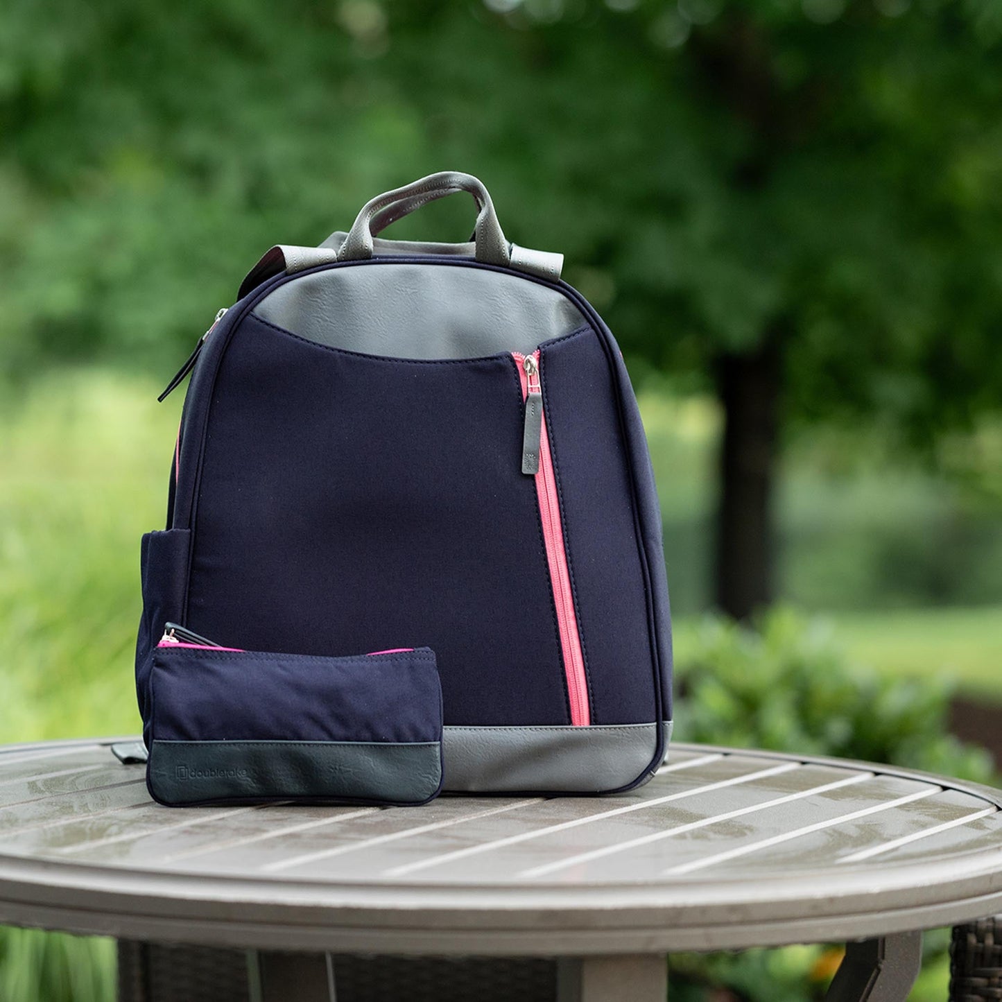 The Doubletake MELBOURNE | Pickleball Backpack & Tote in navy and gray, featuring a pink diagonal zipper, sits on a round outdoor table with its matching pouch, set against lush greenery and blurred trees.