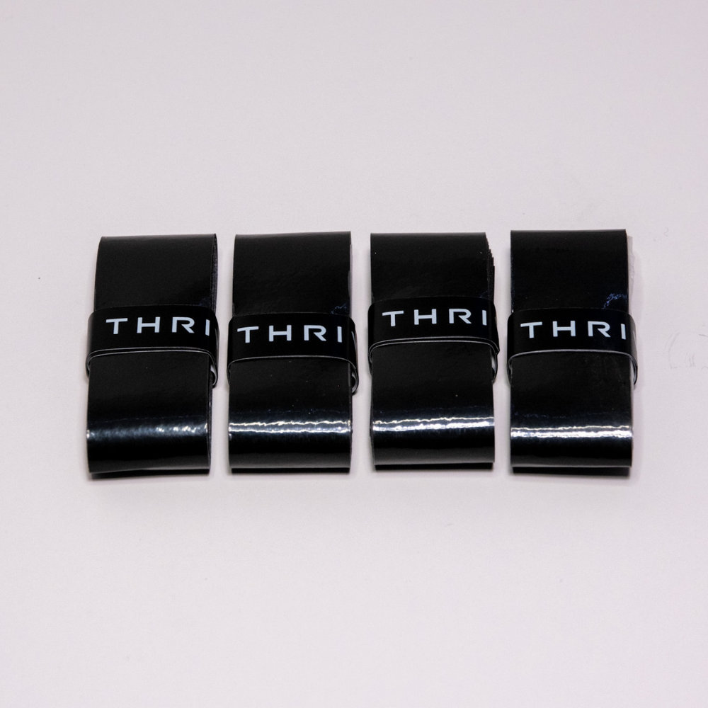 Four identical black Thrive Pickleball Overgrips (Super Tacky) with a glossy finish and super tacky texture are laid side by side on a light surface, each showing a black band with white letters hinting at the brand name.