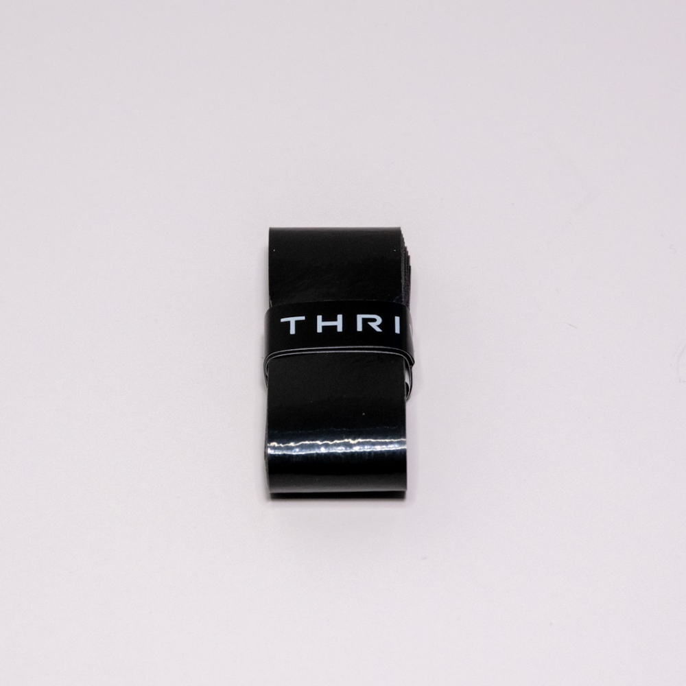 A glossy black rectangular Overgrip by Thrive Pickleball is wrapped with a durable, super tacky black band featuring partial white "THRI" text. The product is centered on a plain, light gray background.